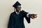 Handsome black man wearing graduation cap and ceremony robe pointing with finger surprised ahead, open mouth amazed expression,