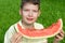 Handsome boy eating a large slice of red watermelon, on a background of green grass