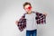 A handsome boy in a plaid shirt, gray shirt and jeans stands on a gray background. A boy in red sunglasses. The boy
