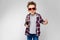 A handsome boy in a plaid shirt, gray shirt and jeans stands on a gray background. A boy wearing sunglasses. The boy