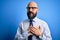 Handsome business bald man with beard wearing elegant tie and glasses over blue background smiling with hands on chest with closed