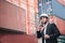Handsome business man wears white hardhat and holding cellphone or smartphone with stacked cargo container. Logistic warehouse