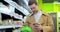 Handsome businessman with shopping trolley using smartphone food checklist walking inside grocery store.