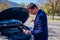 A handsome businessman wearing blue blazer lifting up the hood of his car and checking the oil level on a sunny day parked on a