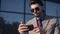 Handsome Bussinessman Wearing Sunglasses and Stylish Suit with Tie Holding his Smartphone Looking into it front of