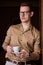 Handsome caucasian stylish guy in brown shirt posing, standing near window with cup of coffee