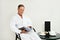 Handsome charming attractive mature European man in white terry bathrobe sitting on armchair and reading magazine, relaxing in