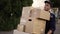 Handsome delivery man in uniform, cap and gloves carrying many heavy cardboard boxes parcels outdoor