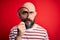 Handsome detective bald man with beard using magnifying glass over red background with a confident expression on smart face