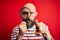 Handsome detective bald man with beard using magnifying glass over red background pointing with finger to the camera and to you,