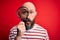 Handsome detective bald man with beard using magnifying glass over red background scared in shock with a surprise face, afraid and