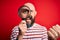 Handsome detective bald man with beard using magnifying glass over red background screaming proud and celebrating victory and