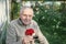 A handsome elderly man with a red rose is sitting on a bench. The old gardener is resting after working in the garden