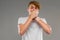 Handsome emotional teenager boy posing in studio against gray, guy in a gray t-shirt covers his mouth with his hands