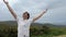 a handsome European man looks at the sky and raises his hands up he is in a white shirt and mountains behind him