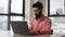 Handsome focused young Indian freelancer male working on laptop computer in office with large windows in daytime.