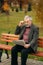 Handsome grandfather with a beautiful beard in a gray jacket sits on a bench in the park and reads a newspaper