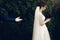 Handsome groom offering his hand to beautiful bride in wedding dress with bouquet, newlywed wife posing in park, proposal concept