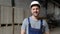 Handsome and happy professional worker in hat charmingly smiling on camera. In the background big warehouse