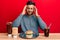 Handsome hispanic man eating a tasty classic burger and soda with hand on head, headache because stress