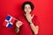 Handsome hispanic man holding dominican republic flag covering mouth with hand, shocked and afraid for mistake