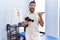 Handsome hispanic man holding therapy massage gun at physiotherapy center pointing thumb up to the side smiling happy with open