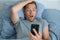 Handsome impressed man reading news on smartphone with shocked expression in bed in the morning. Guy amazed with news