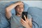 Handsome impressed man reading news on smartphone with upset expression in bed in the morning. Guy amazed with bad news