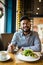 Handsome indian bearded man in checked shirt holding fork eating in cafe and smiling looking at camera