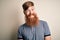 Handsome Irish redhead man with beard and arm tattoo standing over isolated background with a happy and cool smile on face