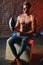 Handsome male bodybuilder with medicine ball sits over brick wall background.