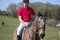 Handsome Male Horse Rider on horseback with white breeches, black boots and red polo shirt in green field with horses in distance