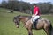 Handsome Male Horse Rider on horseback with white breeches, black boots and red polo shirt in green field with horses in distance