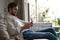 Handsome man in basic t-shirt smiling and holding mobile phone in hands while sitting on couch in living room.