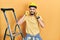 Handsome man with beard by construction stairs wearing hardhat covering ears with fingers with annoyed expression for the noise of