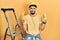 Handsome man with beard by construction stairs wearing hardhat gesturing finger crossed smiling with hope and eyes closed