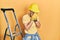 Handsome man with beard by construction stairs wearing hardhat with sad expression covering face with hands while crying