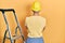 Handsome man with beard by construction stairs wearing hardhat standing backwards looking away with crossed arms