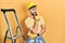 Handsome man with beard by construction stairs wearing hardhat thinking worried about a question, concerned and nervous with hand