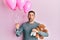 Handsome man with beard expecting a baby girl holding balloons, shoes and teddy bear making fish face with mouth and squinting