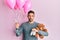 Handsome man with beard expecting a baby girl holding balloons, shoes and teddy bear relaxed with serious expression on face