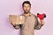 Handsome man with beard holding packing tape holding cardboard making fish face with mouth and squinting eyes, crazy and comical