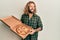 Handsome man with beard and long hair holding italian pizza angry and mad screaming frustrated and furious, shouting with anger