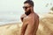 Handsome Man with beard, In Sunglasses Sunbathing With Sunscreen Lotion Body In Summer. Male Fitness Model Tanning Using Solar Blo