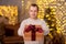 Handsome man giving gift box over christmas background with Christmas tree and led lights, focus on present