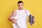 Handsome man with his small newborn daughter posing isolated over yellow background and holding iron in hands, being confused,