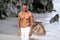Handsome man shirtless and in white pants walking along on sandy beach. Guy with sexy fitness body.