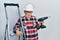 Handsome mature handyman holding screwdriver wearing hardhat by construction stairs depressed and worry for distress, crying angry