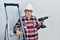 Handsome mature handyman holding screwdriver wearing hardhat by construction stairs in shock face, looking skeptical and