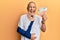 Handsome mature injured man wearing cervical collar and sling holding insurance euros sticking tongue out happy with funny